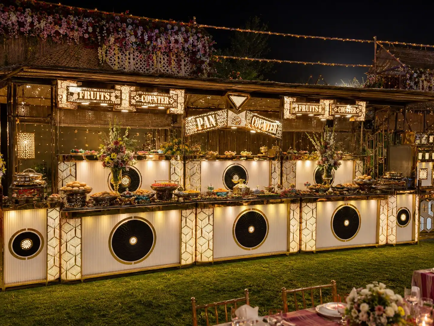 Illuminated fruit and pan buffet counters on a lawn at night